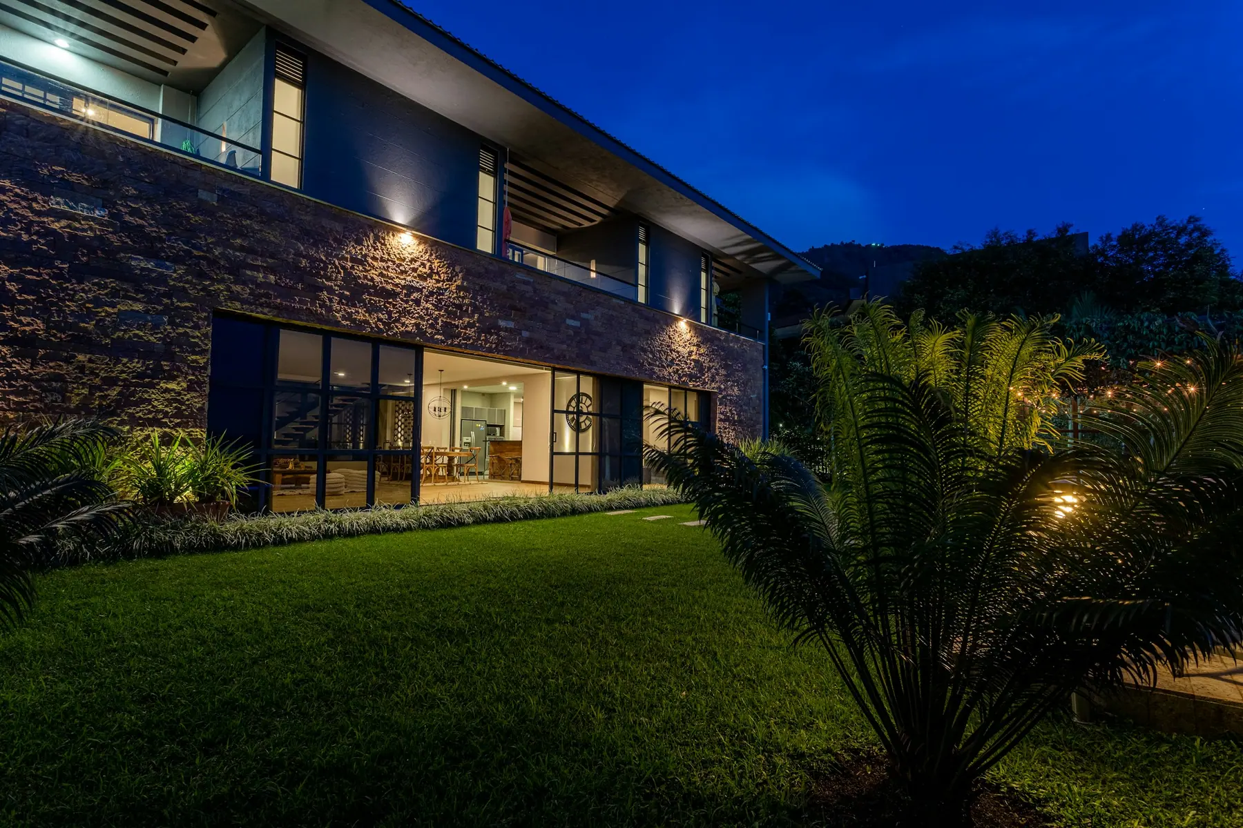 Fachada de pedra e vidro da Casa Serra do Cipó iluminada ao anoitecer, com gramado em primeiro plano