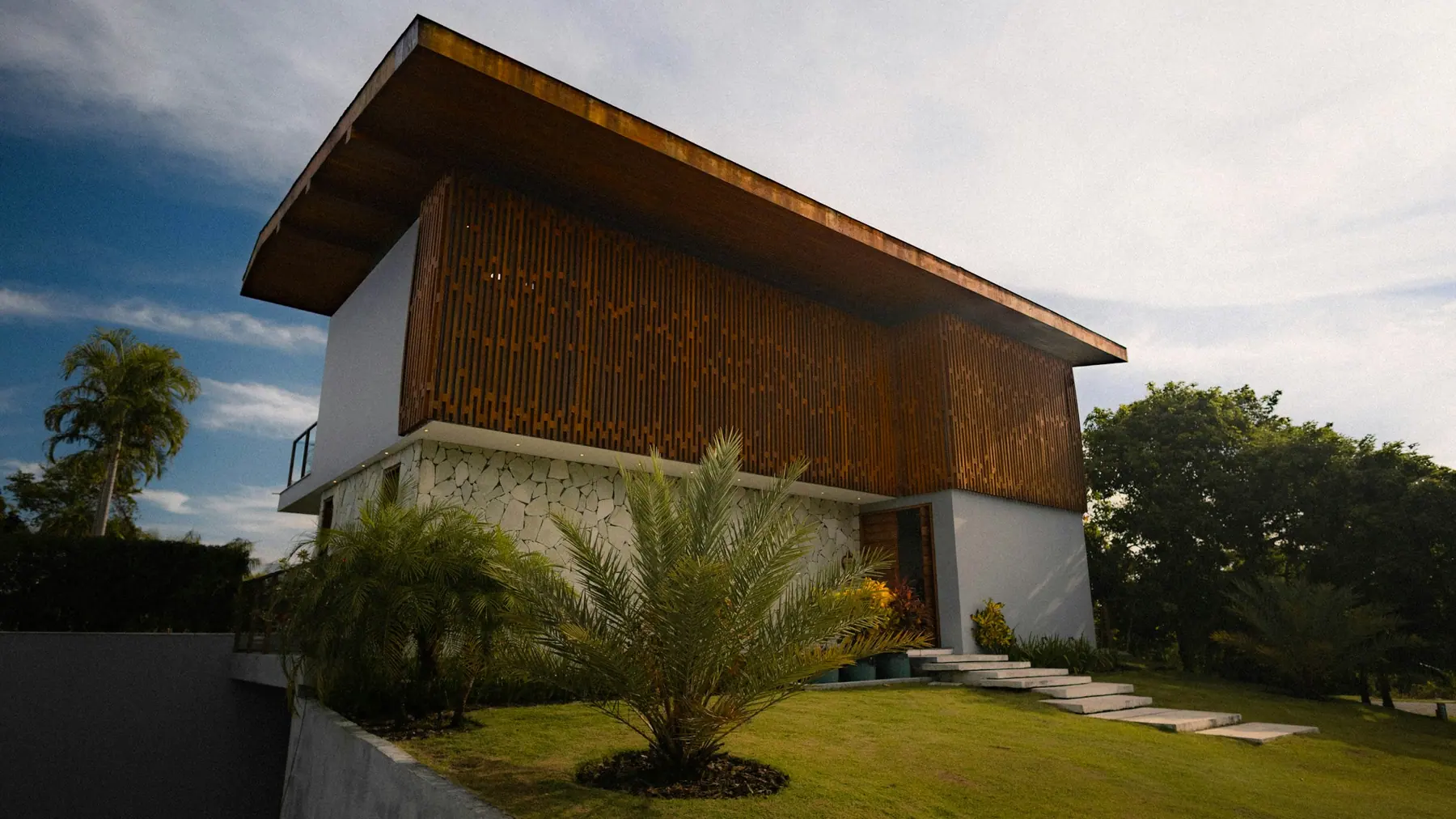 Casa com fachada de ripas de madeira, base de pedra e jardim com palmeiras